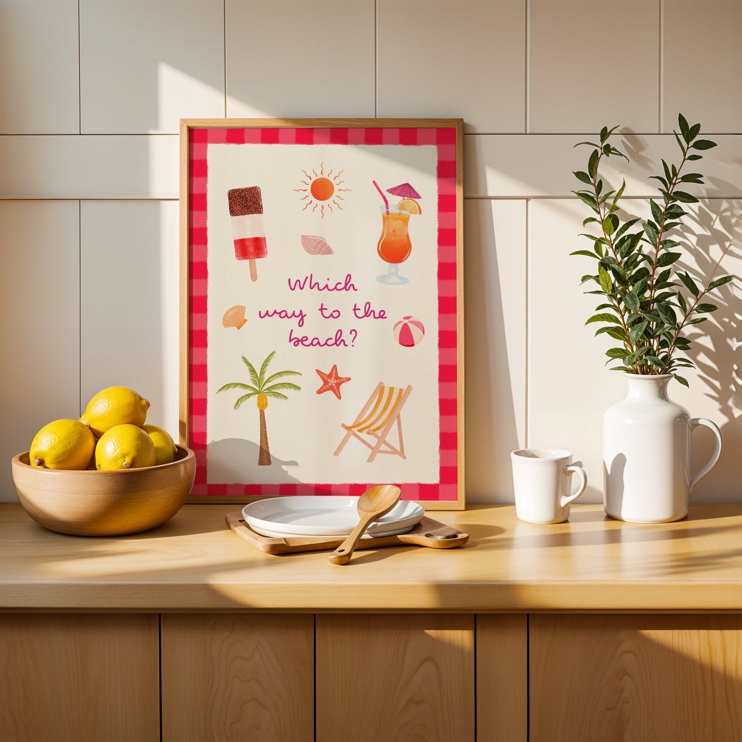 A framed summer-themed art print with the text 'Which way to the beach?' and beach illustrations, styled on a wooden counter next to lemons, a mug, and a potted plant against a tiled wall.