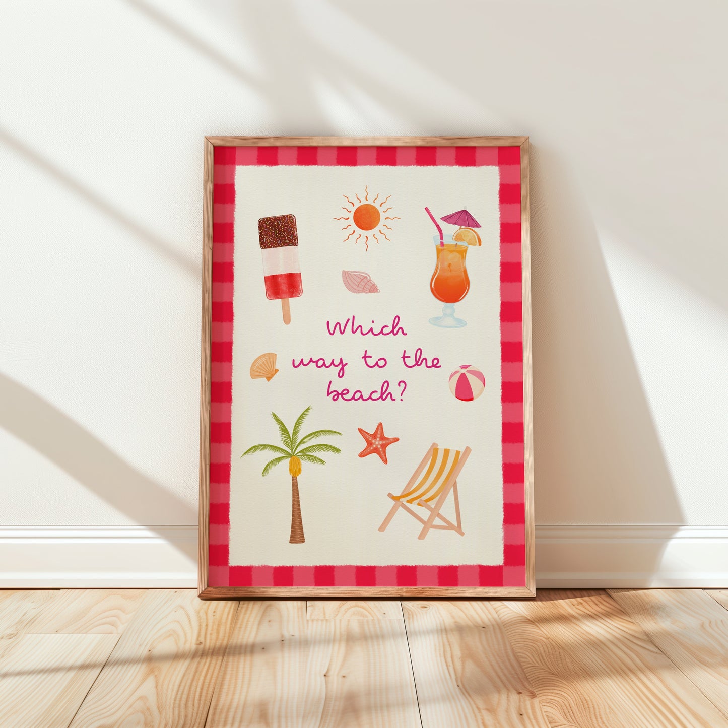 A framed art print leaning against a white wall on a light wood floor. The print features a red and pink striped border and a white textured background with summer beach-themed illustrations including a palm tree, deck chair, cocktail, and sun, with the text 'Which way to the beach?'.