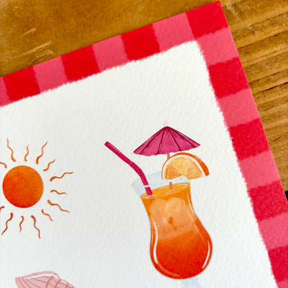 A close-up of a printed art print showing the texture of the paper. Featuring a hand-drawn tropical drink with a pink umbrella and straw, a sun, and a red-and-pink checkered border, placed on a wooden surface.
