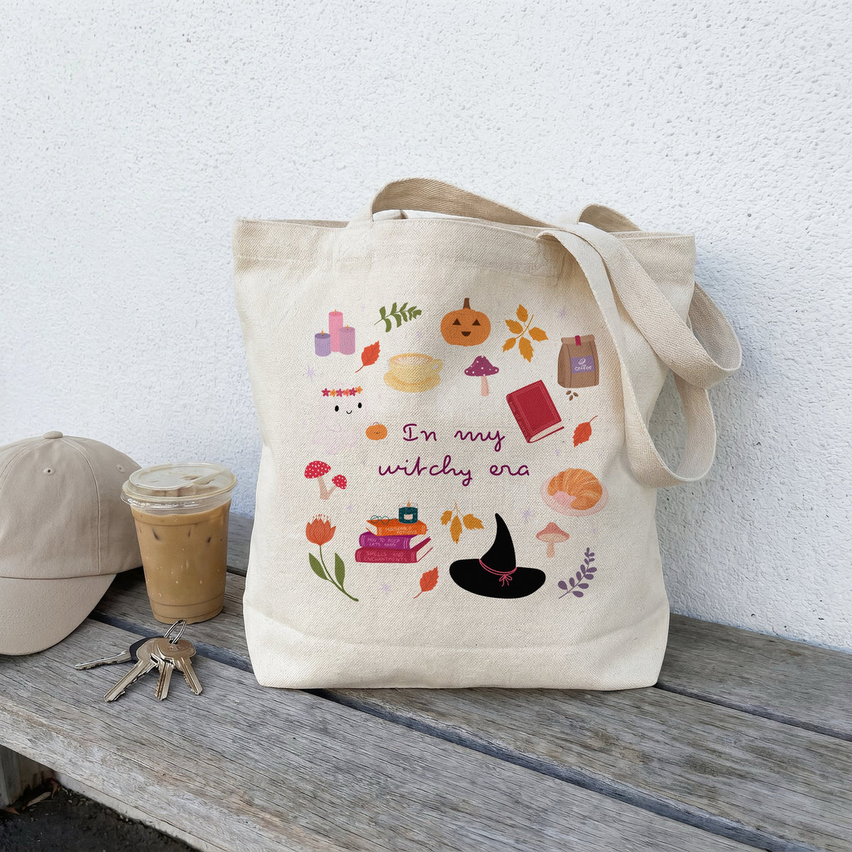 A beige canvas tote bag with a colourful autumn witch-themed graphic print sits on a weathered wooden bench next to a beige baseball cap, keys, and an iced coffee against a white stucco wall.