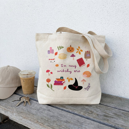 A beige canvas tote bag with a colourful autumn witch-themed graphic print sits on a weathered wooden bench next to a beige baseball cap, keys, and an iced coffee against a white stucco wall.