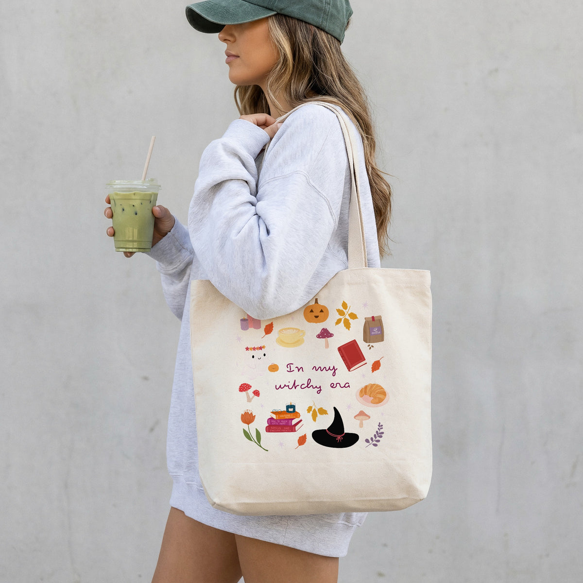 A woman wearing a green cap and a light grey sweatshirt carries a cream canvas tote bag with a colourful autumn witch-themed illustration and the text 'in my witchy era'. She is holding an iced coffee against a neutral grey wall.