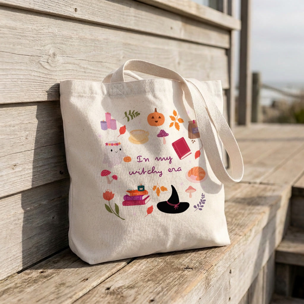An off-white canvas tote bag with a 'Witchy Era' Halloween graphic print, leaning against a rustic wooden boardwalk railing.