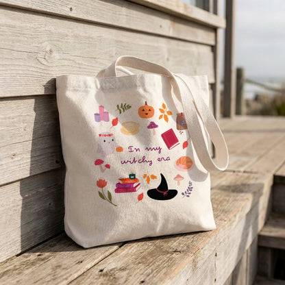 An off-white canvas tote bag with a 'Witchy Era' Halloween graphic print, leaning against a rustic wooden boardwalk railing.
