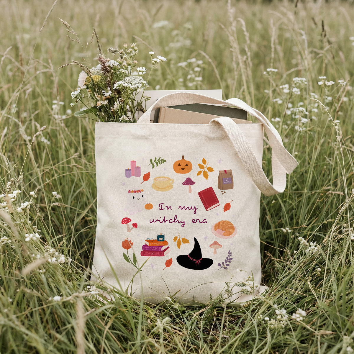 A cream canvas tote bag with a whimsical autumn witch-themed print, sitting in a grassy field with wildflowers. The bag features illustrations of a witch's hat, books, a pumpkin, mushrooms, and the text 'In my witchy era'.