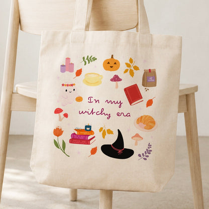 A cream canvas tote bag with a colourful autumn witch illustration and the text 'In my witchy era' hanging from a light wood chair.