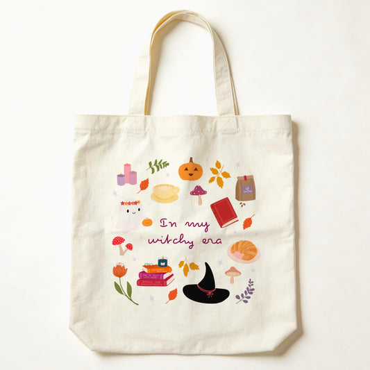 An off-white canvas tote bag with a cozy autumn witchy theme, featuring a central graphic with illustrations of a witch's hat, potion bottles, a pumpkin, books, and the text 'In my witchy era'.