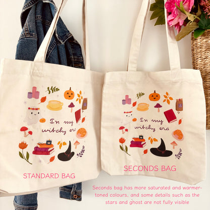 Two canvas tote bags displayed side by side against a white wall, with a denim jacket hanging on the left and flowers in a basket on the upper right. The left bag is labelled 'STANDARD BAG' and the right bag 'SECONDS BAG'; both are off-white with a whimsical witch-themed illustration including pumpkins, books, mushrooms, and a black hat, plus pink text reading 'In my witchy era'.