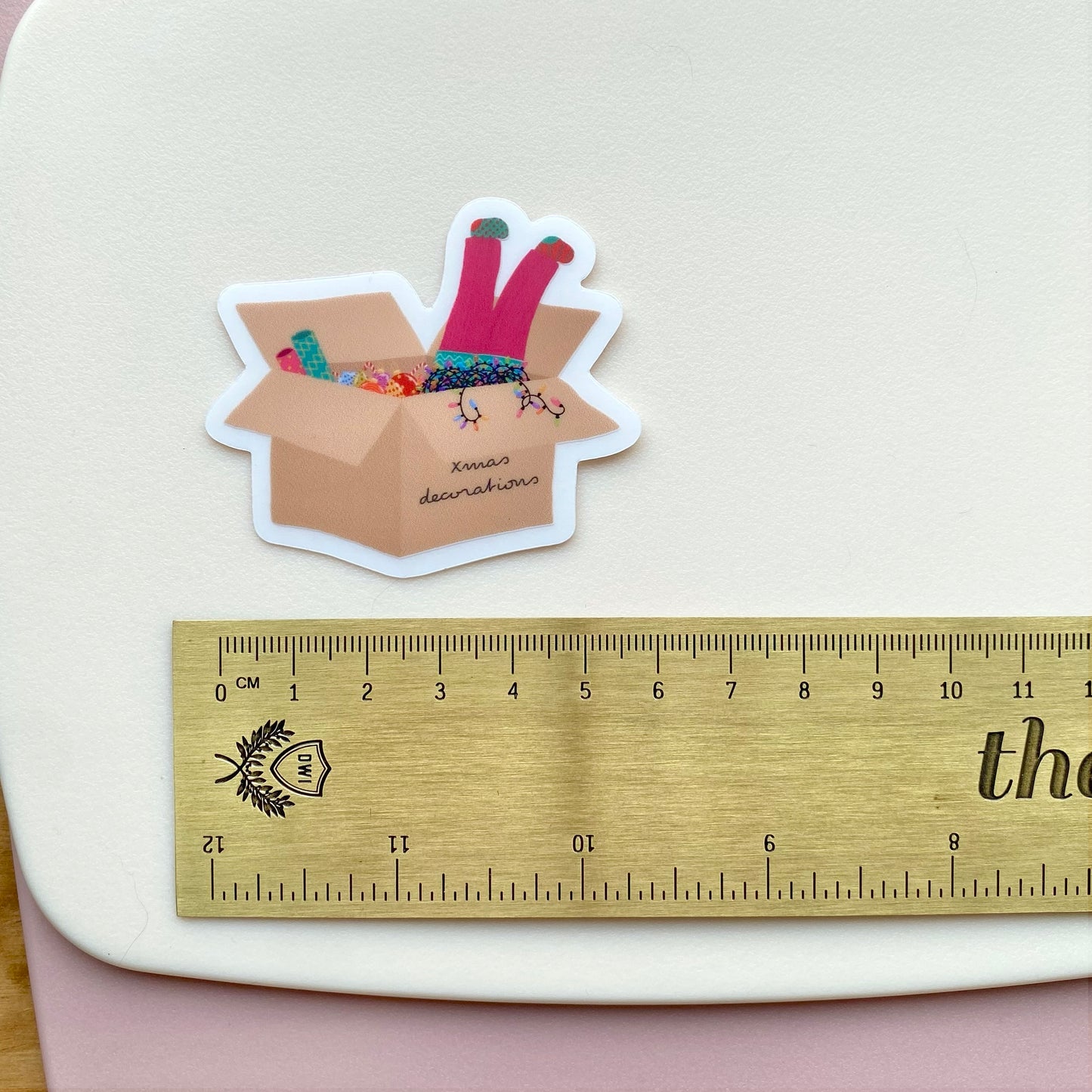 Sticker of a Christmas decorations box with colourful baubles and lights and a girl stuck head first inside, next to a ruler on a light background