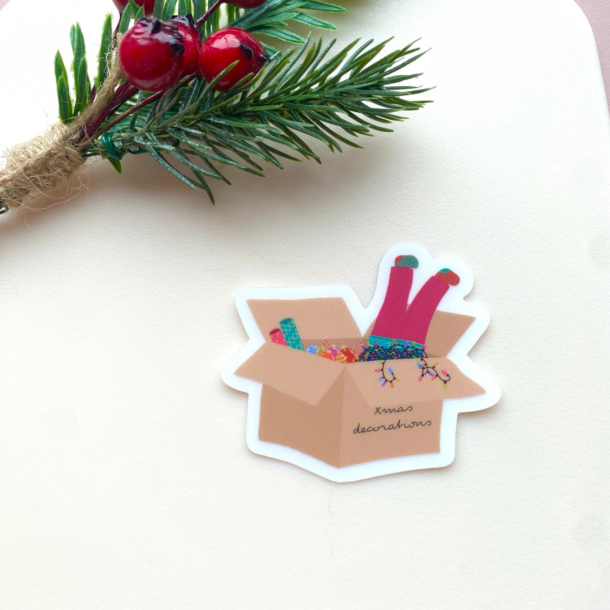 Sticker of a cardboard box with Christmas decorations and a girls legs dangling out with pink trousers and mismatched socks, on a white background with festive foliage and berries.