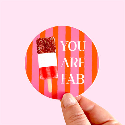 A hand holding a round sticker with a pink and orange striped background, a popsicle illustration, and the text 'YOU ARE FAB' in white. The background of the photo is a solid light pink.