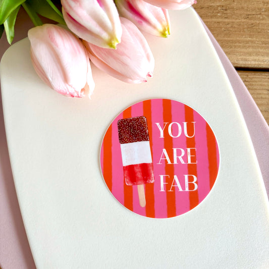 A round sticker with a pink and orange striped background, featuring a red and white popsicle and the text 'YOU ARE FAB' in white. The sticker is placed on a white surface next to pink tulips.