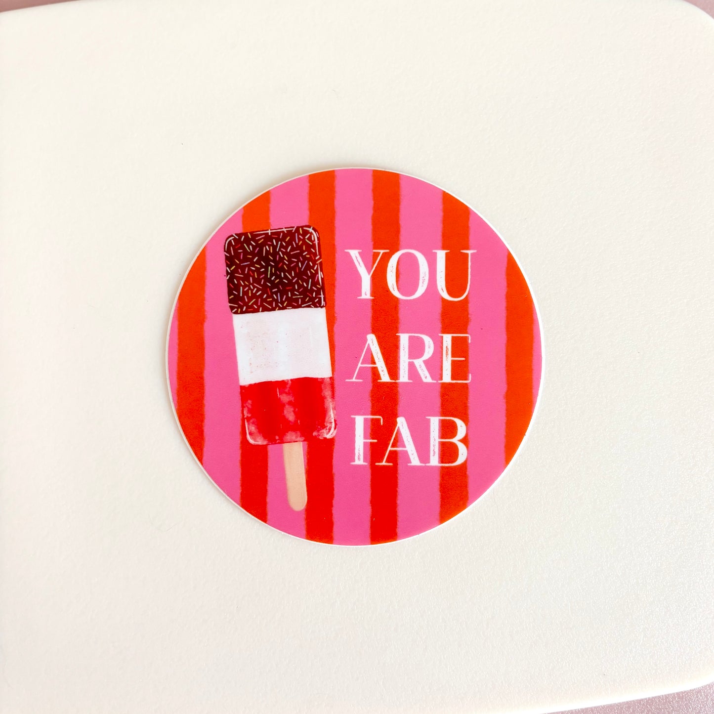 A round sticker with a pink and red striped background, featuring a chocolate-dipped popsicle and the text 'YOU ARE FAB' in white, shown on a light cream surface.
