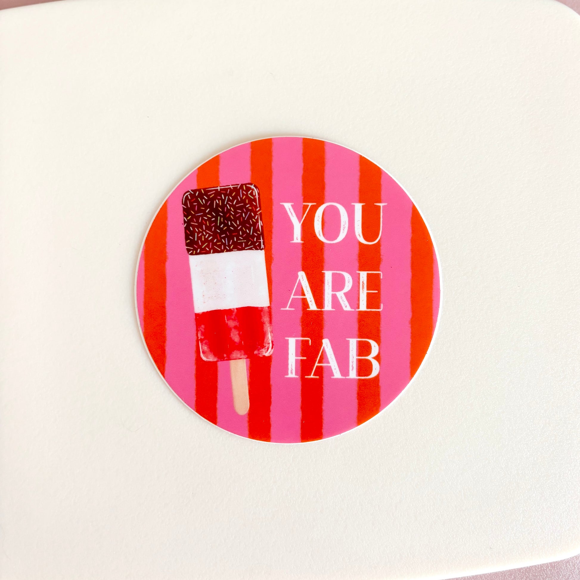 A round sticker with a pink and red striped background, featuring a chocolate-dipped popsicle and the text 'YOU ARE FAB' in white, shown on a light cream surface.