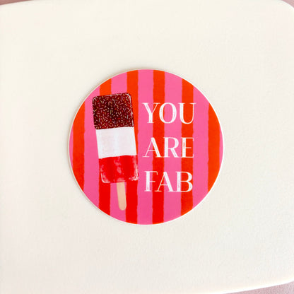 A round sticker with a pink and red striped background, featuring a chocolate-dipped popsicle and the text 'YOU ARE FAB' in white, shown on a light cream surface.