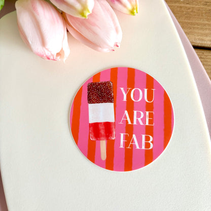 A round sticker with a pink and orange striped background, featuring a popsicle graphic and the text 'YOU ARE FAB' in white. The sticker is displayed on a white card with pink tulips in the background.