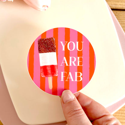 A hand holds a round sticker against a cream-colored card. The sticker features a pink and orange striped background, a watercolor-style popsicle with red, white, and brown layers, and the text 'YOU ARE FAB' in white.