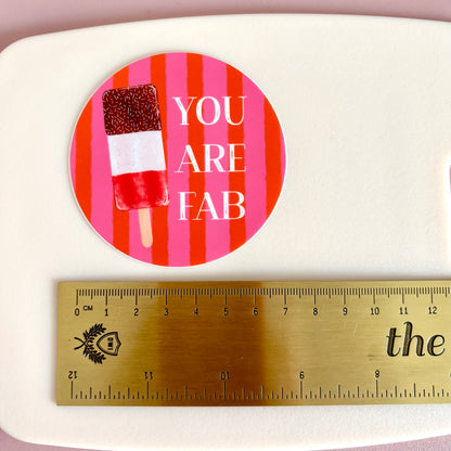 A round sticker with a pink and red striped background, a popsicle graphic, and the text 'YOU ARE FAB' in white, shown above a gold metal ruler on a white surface.