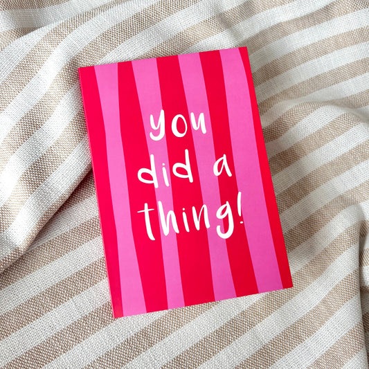 A pink striped greeting card with the white handwritten text 'you did a thing!' resting on a beige and white striped fabric background.