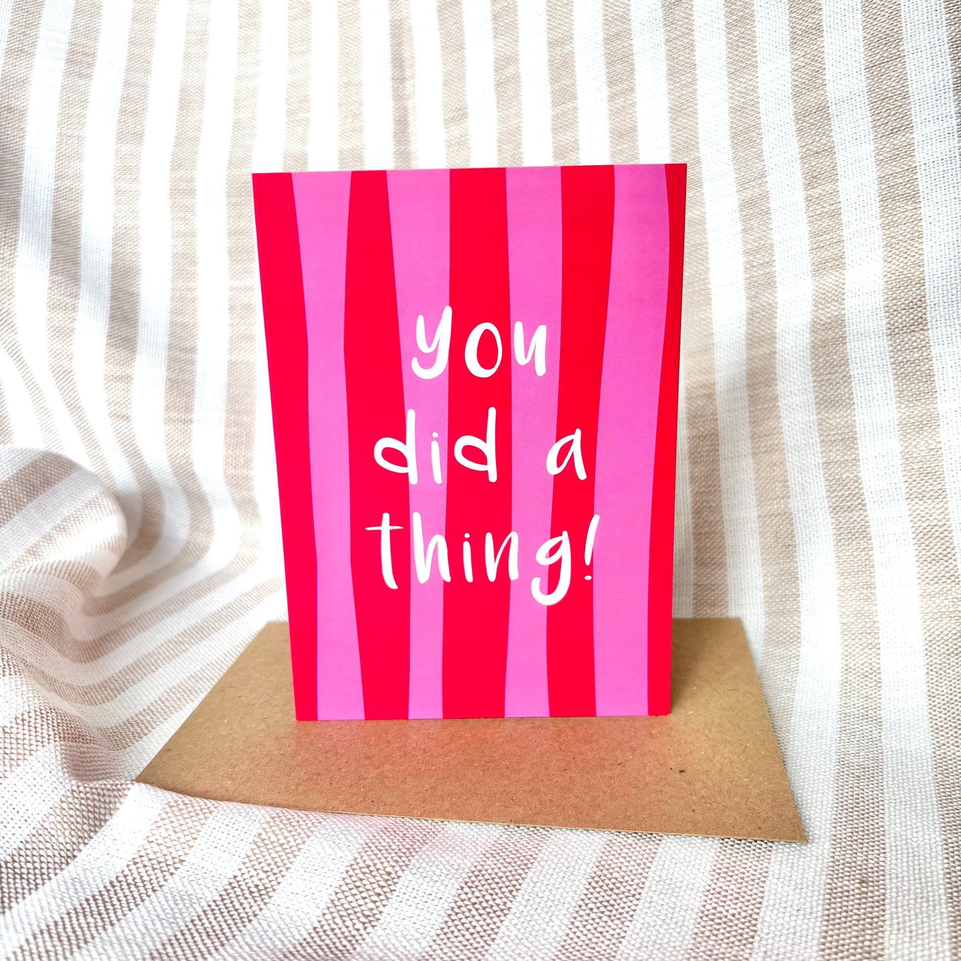 A pink and red vertically striped greeting card with the white text 'you did a thing!' stands upright on a brown envelope against a beige and white striped fabric background.