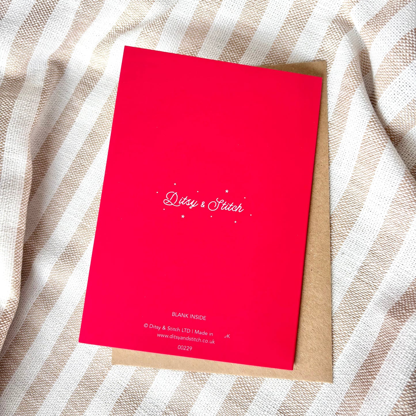 The back of a bright pink greeting card with a white brand logo reading 'Ditsy & Stitch', placed on top of a brown kraft envelope over a beige and white striped fabric background.