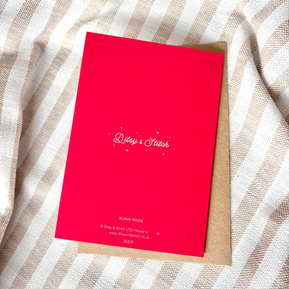 The back of a bright pink greeting card with a white brand logo reading 'Ditsy & Stitch', placed on top of a brown kraft envelope over a beige and white striped fabric background.