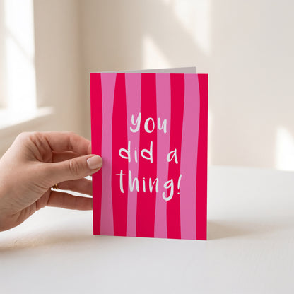 A hand holds a pink striped greeting card that reads 'you did a thing!' in white script against a softly lit indoor background.