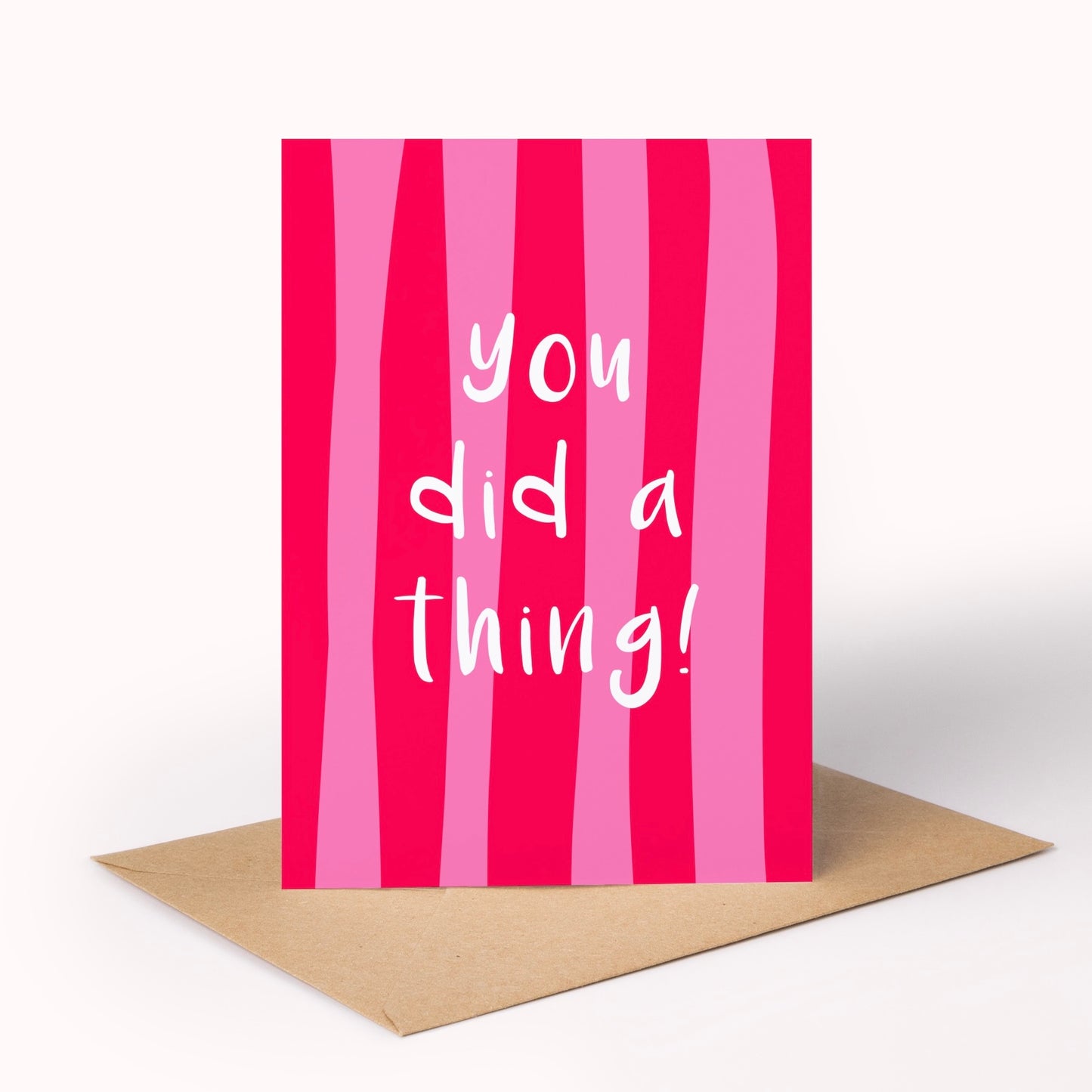 A pink and magenta vertically striped greeting card with the white handwritten text 'you did a thing!' standing upright, placed on a brown kraft envelope against a clean white background.