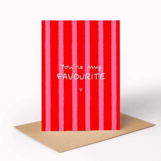 A red and pink vertically striped greeting card with the white text 'You're my FAVOURITE' and a small heart, shown standing upright on a brown envelope against a white background.