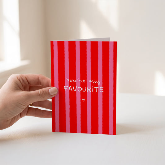 A hand holds a red and pink vertically striped greeting card that reads 'You're my FAVOURITE' in white script and block lettering, set against a bright, softly lit indoor background.