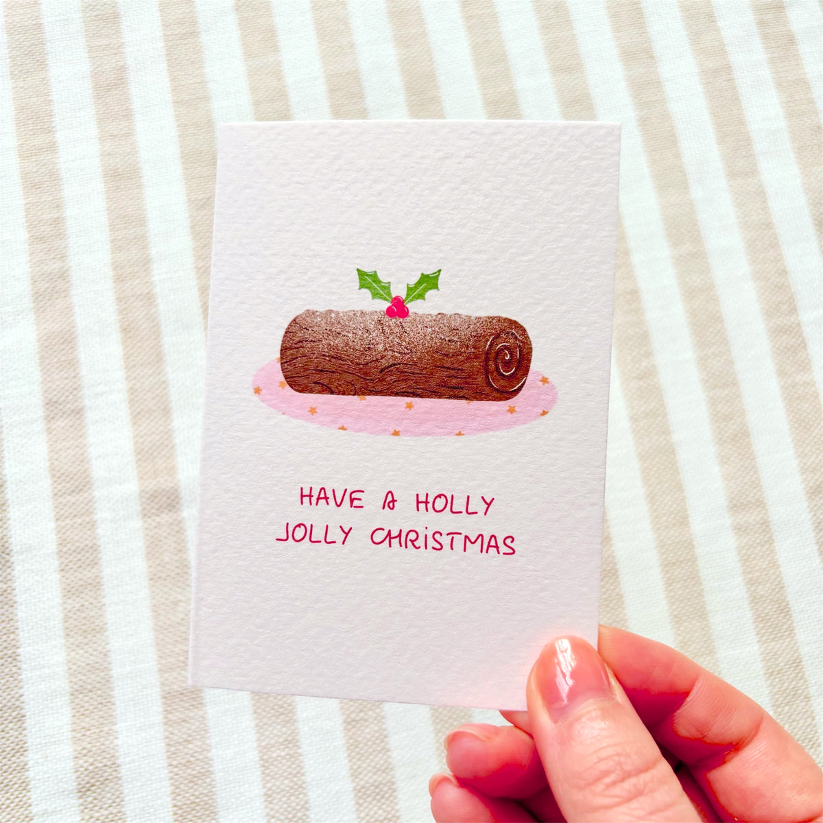 A hand holds a white greeting card featuring a watercolor illustration of a Christmas yule log with holly leaves and a pink plate, with the text 'HAVE A HOLLY JOLLY CHRISTMAS' below. The background is a beige and white striped fabric.
