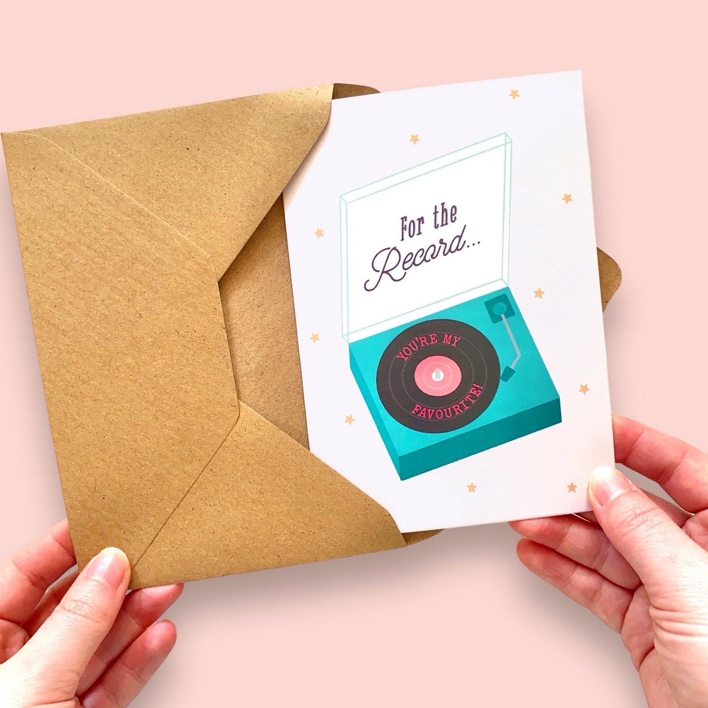 Greeting card with record player design  and 'For the record... you're my favourite' message held up by hand with an envelope against a pink background.