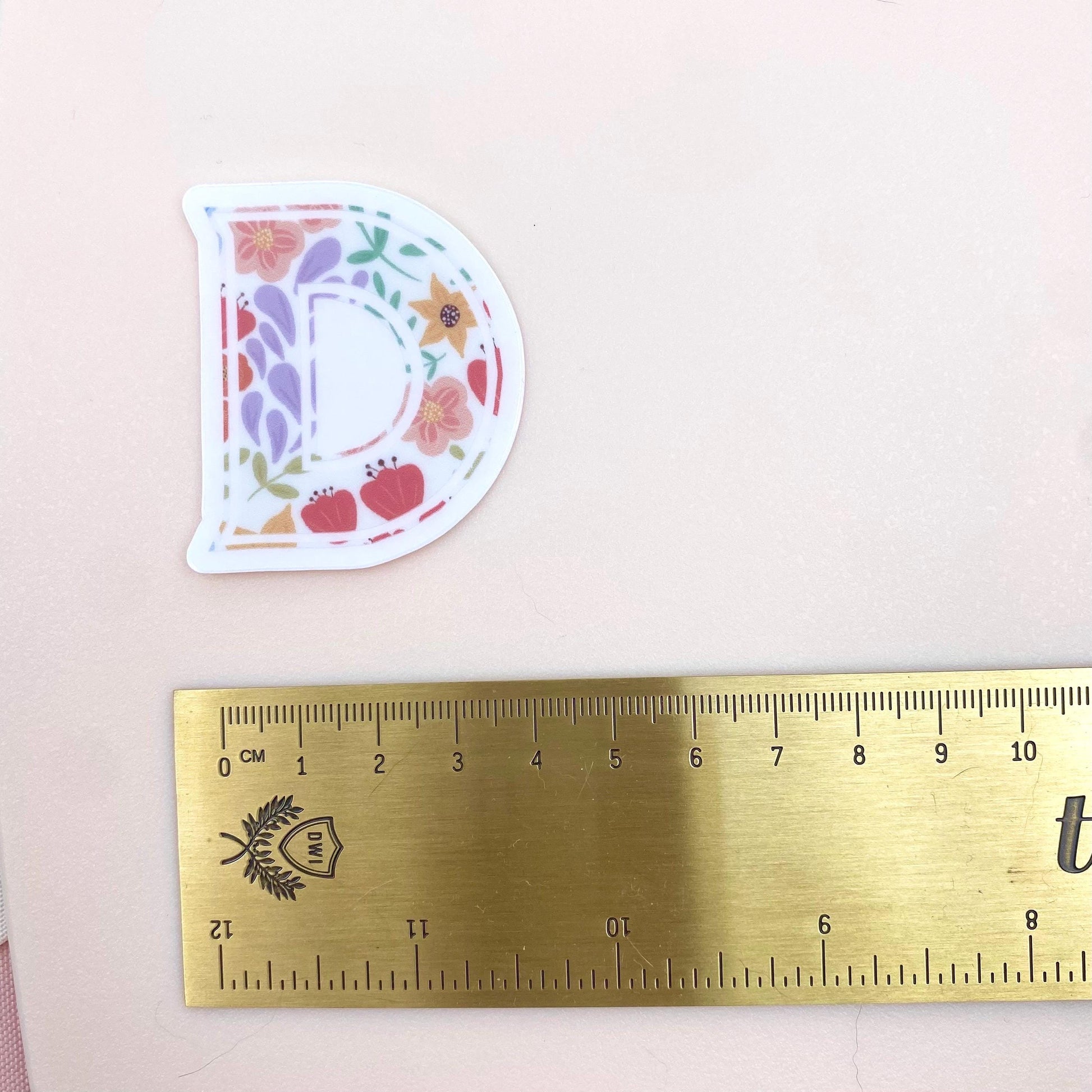 Floral letter 'D' sticker next to a ruler on a pink background
