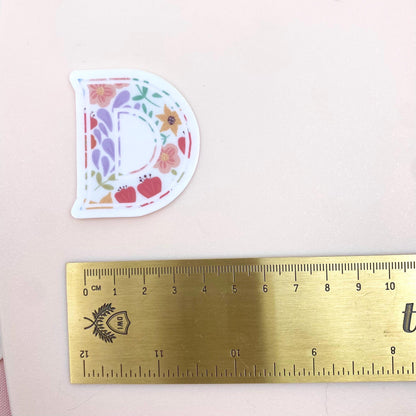 Floral letter 'D' sticker next to a ruler on a pink background