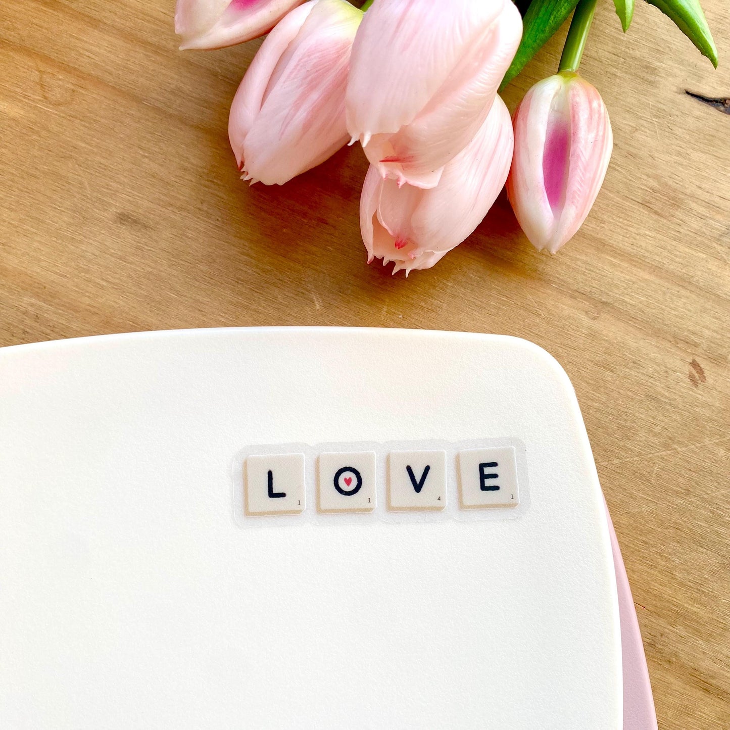 A sticker with Scrabble tiles spelling 'LOVE' on a white surface with pink tulips in the background.