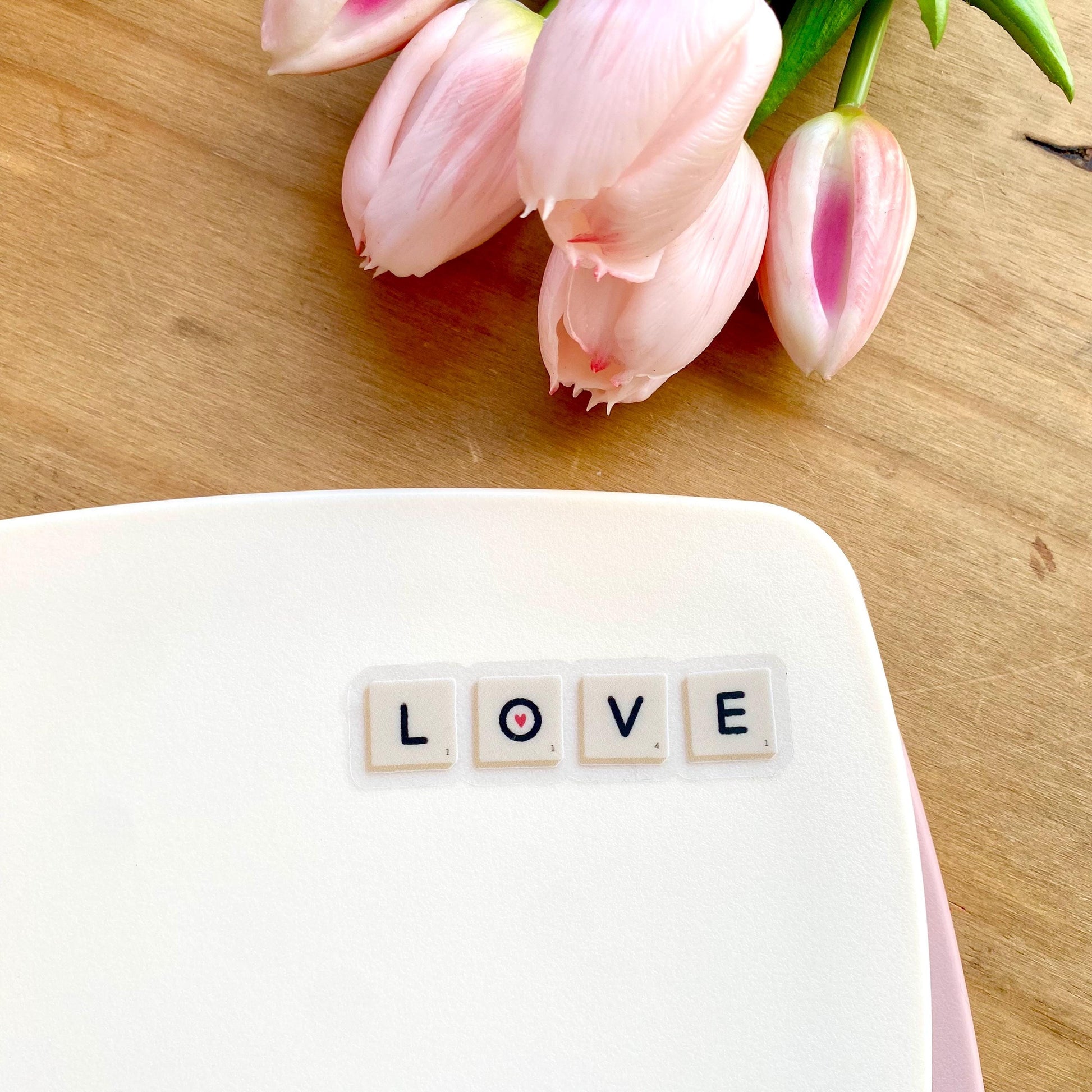 A sticker with Scrabble tiles spelling 'LOVE' on a white surface with pink tulips in the background.