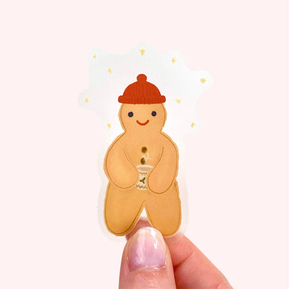 Gingerbread man sticker with a red hat held between fingers on a white background