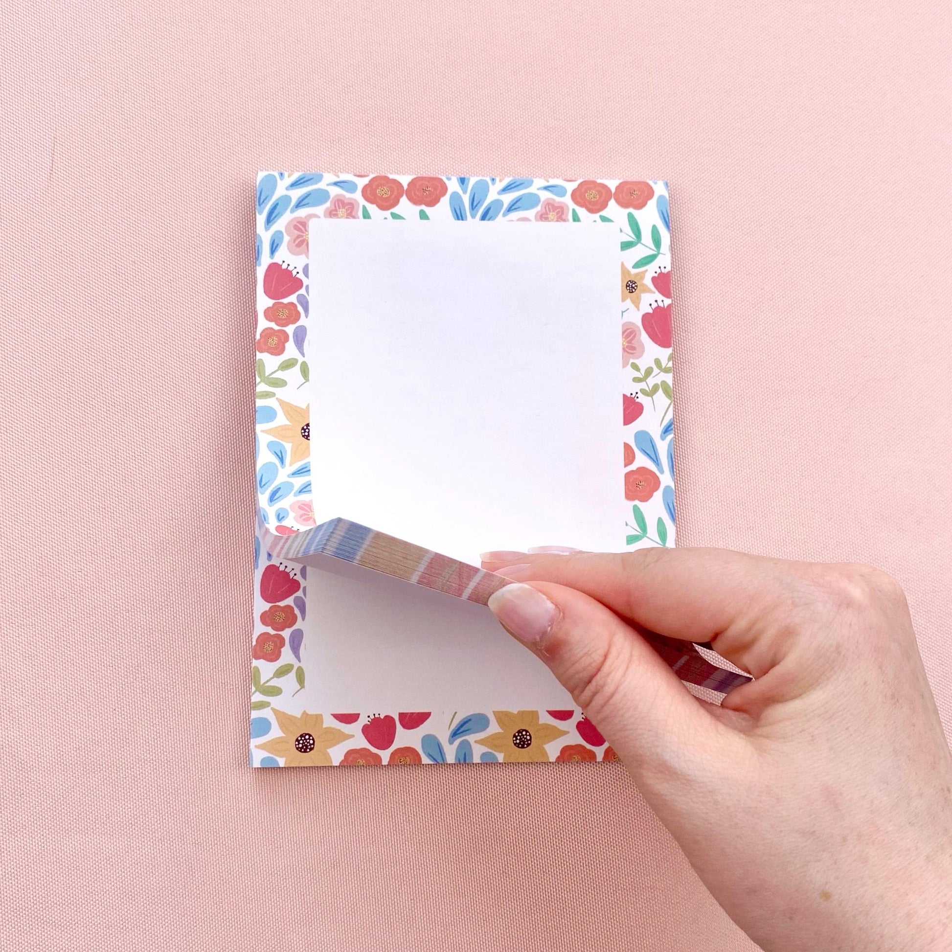 Floral-patterned notepad with a hand lifting a page against a pink background