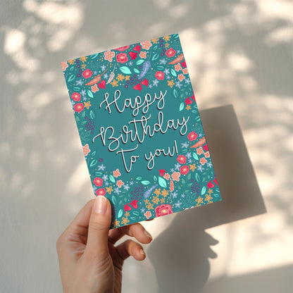 Hand holding a colorful birthday card with floral design and 'Happy Birthday To You!' text.
