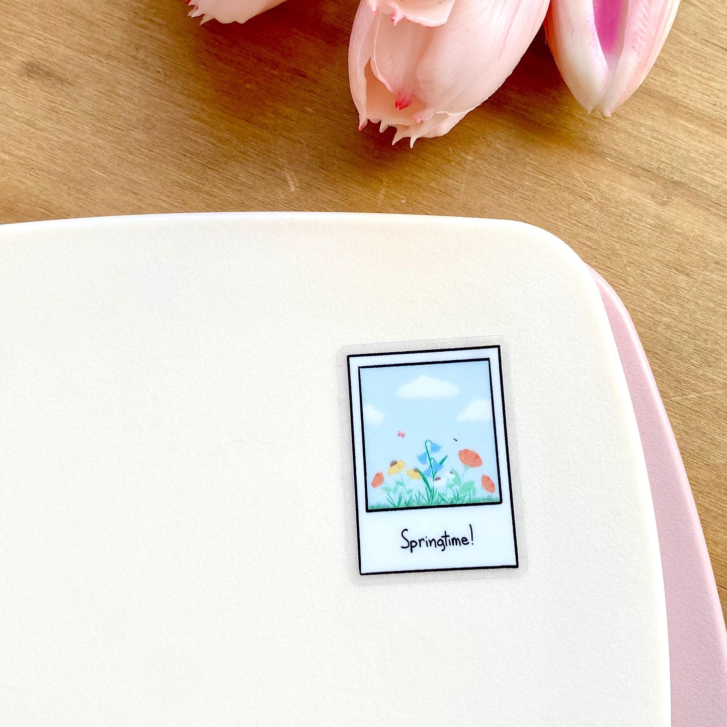 Sticker with 'Springtime!' polaroid design on a white surface with pink tulips in the background.