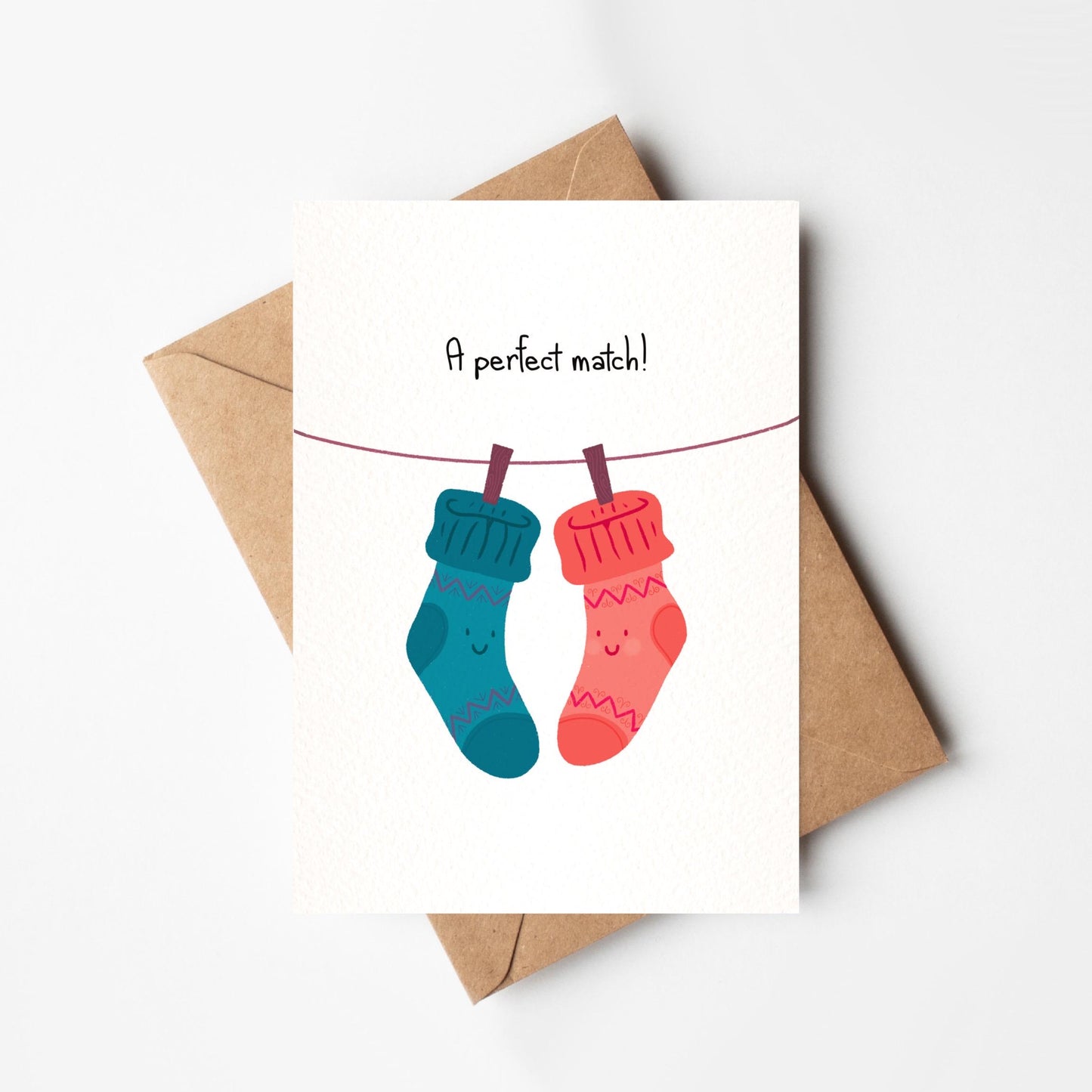 Greeting card with two socks on a line and 'A perfect match!' text, next to a brown envelope on a white background.