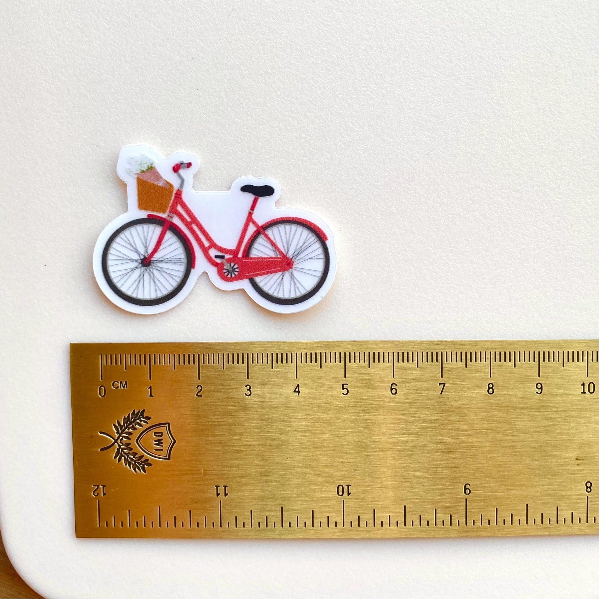 Pink bicycle sticker against a cream background with a gold ruler for scale.
