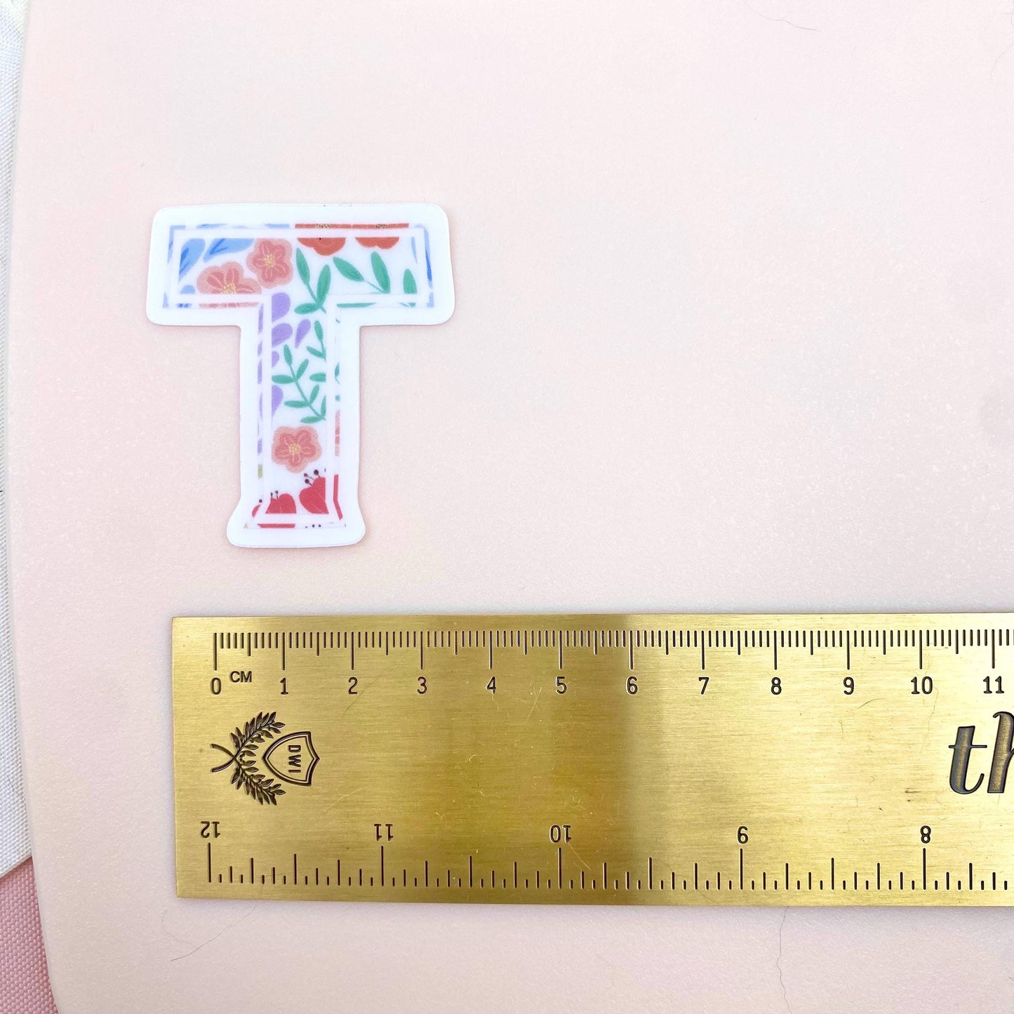 Floral letter 'T' sticker next to a ruler on a pink background