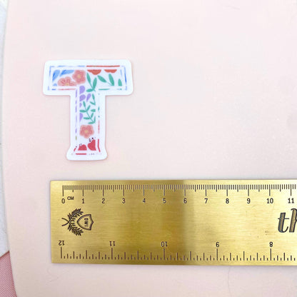 Floral letter 'T' sticker next to a ruler on a pink background