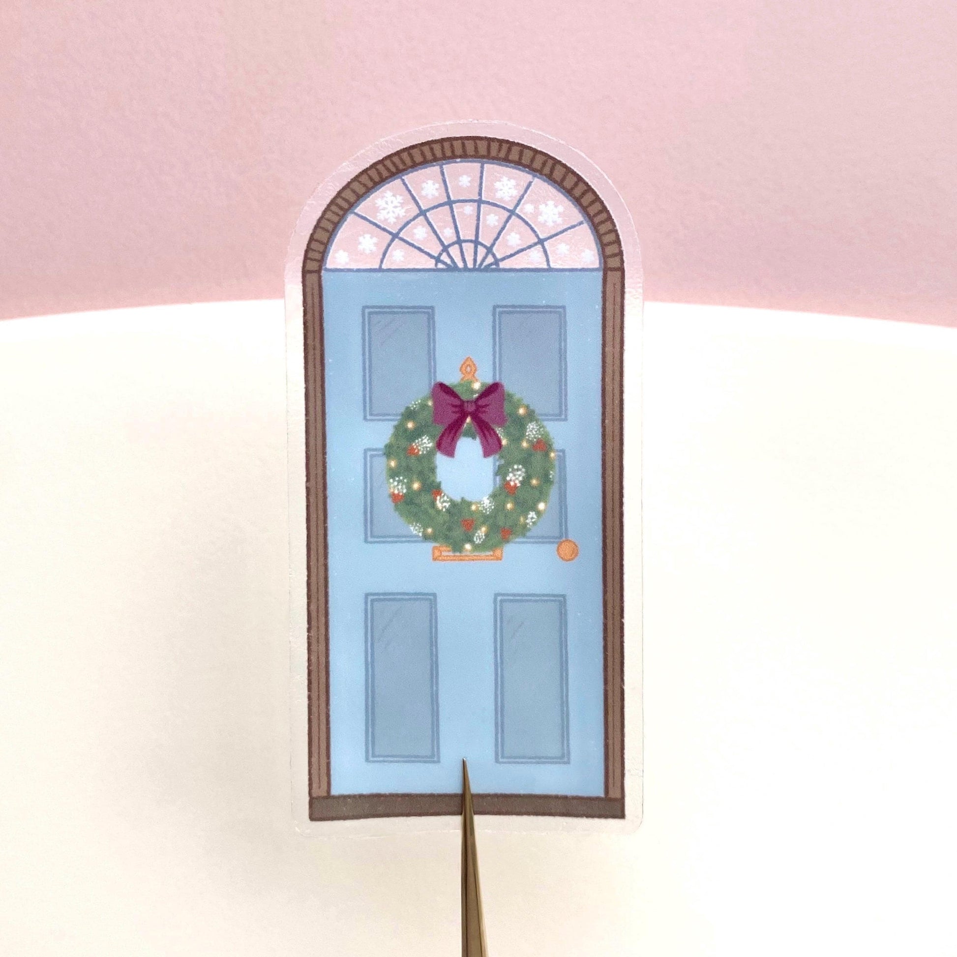 A sticker with a blue door decorated for Christmas held against a pink and cream background.