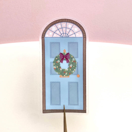 A sticker with a blue door decorated for Christmas held against a pink and cream background.