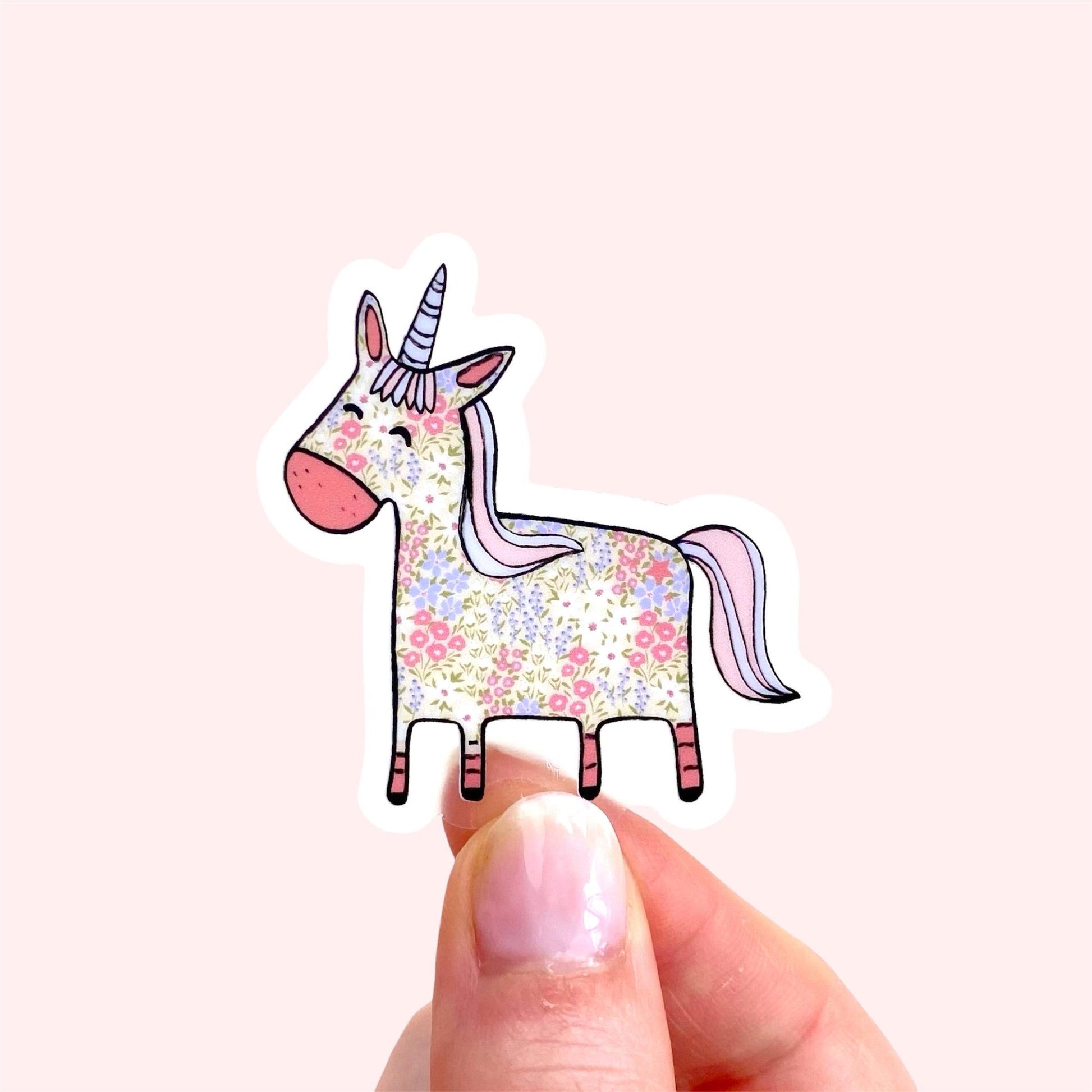 Hand holding a colorful unicorn sticker with floral patterns on a light pink background