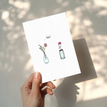Hand holding a card with illustrations of vases and flowers on a light background