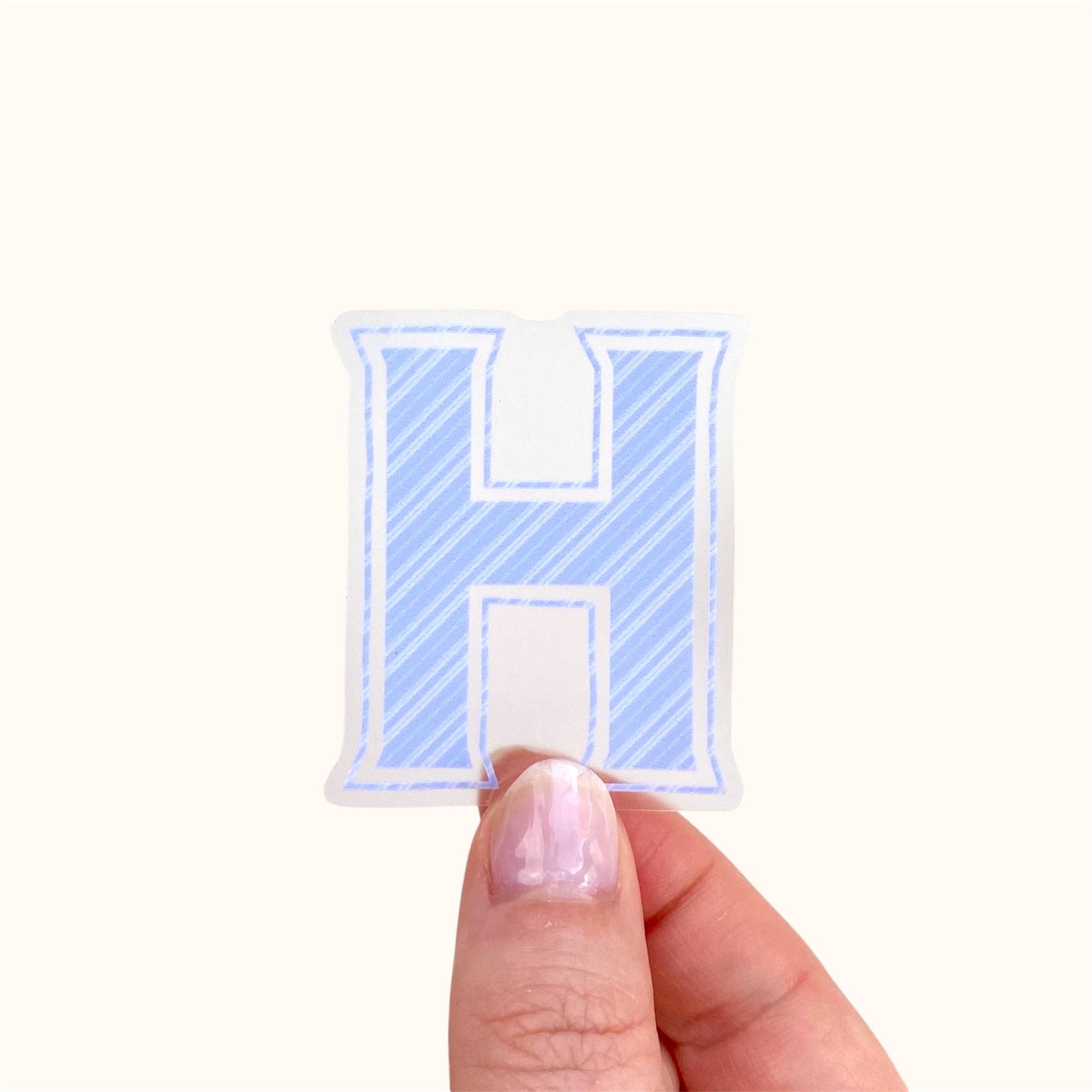 Hand holding a blue striped letter 'H' sticker against a white background