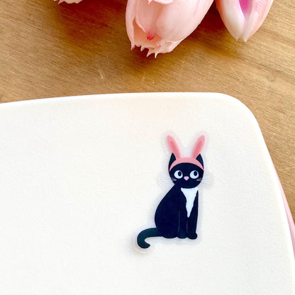 Sticker of a black cat with pink bunny ears on a white surface.